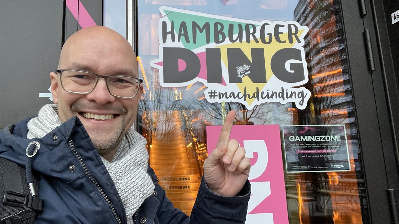 Person lächelt vor dem Eingang des ‚Hamburger Ding‘, mit einem Schild im Hintergrund, das den Slogan ‚#machdeinding‘ zeigt. Die Szene strahlt eine freundliche und inspirierende Atmosphäre aus.