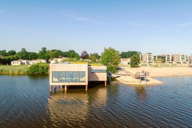 Eine moderne Seelodge mit großen Glasfronten und Holzverkleidung, die direkt über dem Wasser gebaut ist. Im Hintergrund ist ein weitläufiger Strandbereich mit Personen sowie eine Ferienanlage mit modernen Häusern zu sehen. Das Gebäude ist über einen Steg erreichbar.