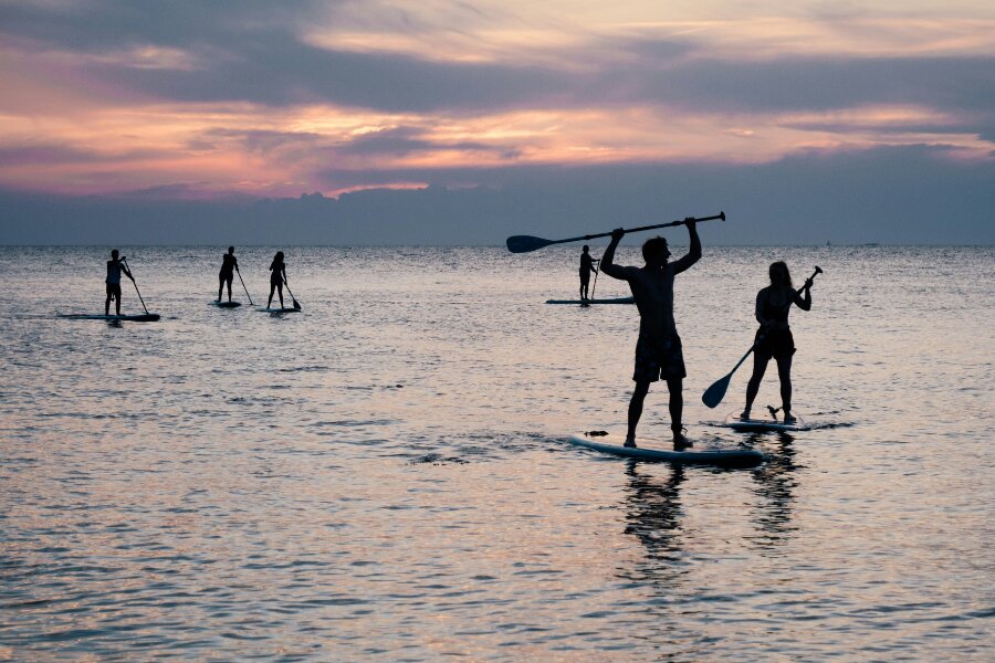 Gruppe von Menschen beim Stand-Up-Paddling auf einem ruhigen Meer bei Sonnenuntergang. Die Silhouetten spiegeln sich im sanft schimmernden Wasser, während der Himmel in warmen Rosa- und Orangetönen leuchtet.