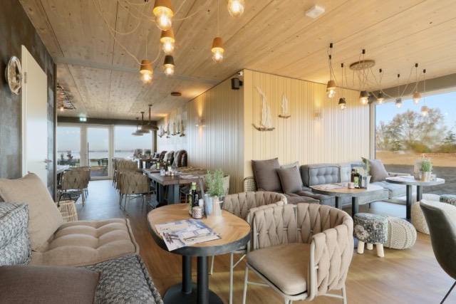 Ein stilvoll eingerichtetes Restaurant mit einem skandinavisch inspirierten Design, gemütlichen Sesseln, Holztischen und dezenten maritimen Dekorationen an der Wand. Große Fenster lassen viel Licht in den Raum und bieten eine Aussicht auf die Umgebung.