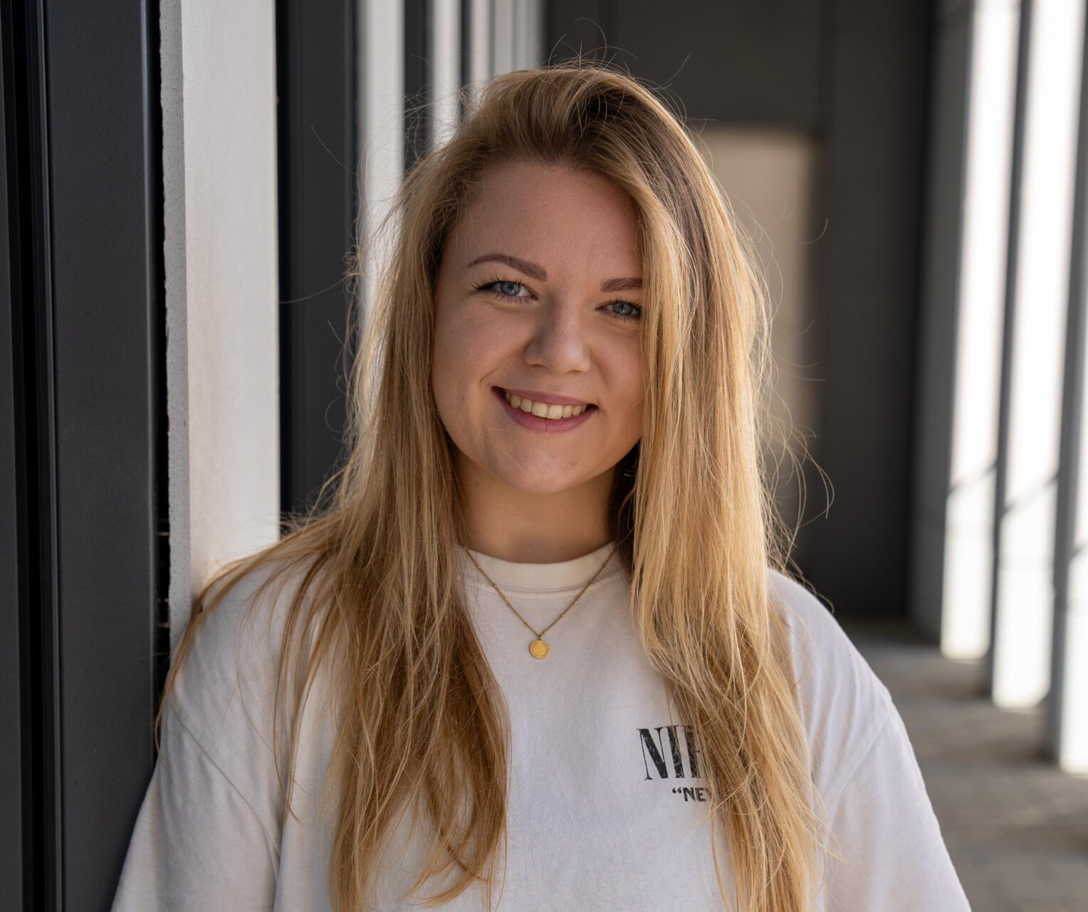 Porträt einer jungen Frau mit langen blonden Haaren, die lächelnd an einer modernen Gebäudewand lehnt. Sie trägt ein weißes Shirt und eine goldene Halskette.