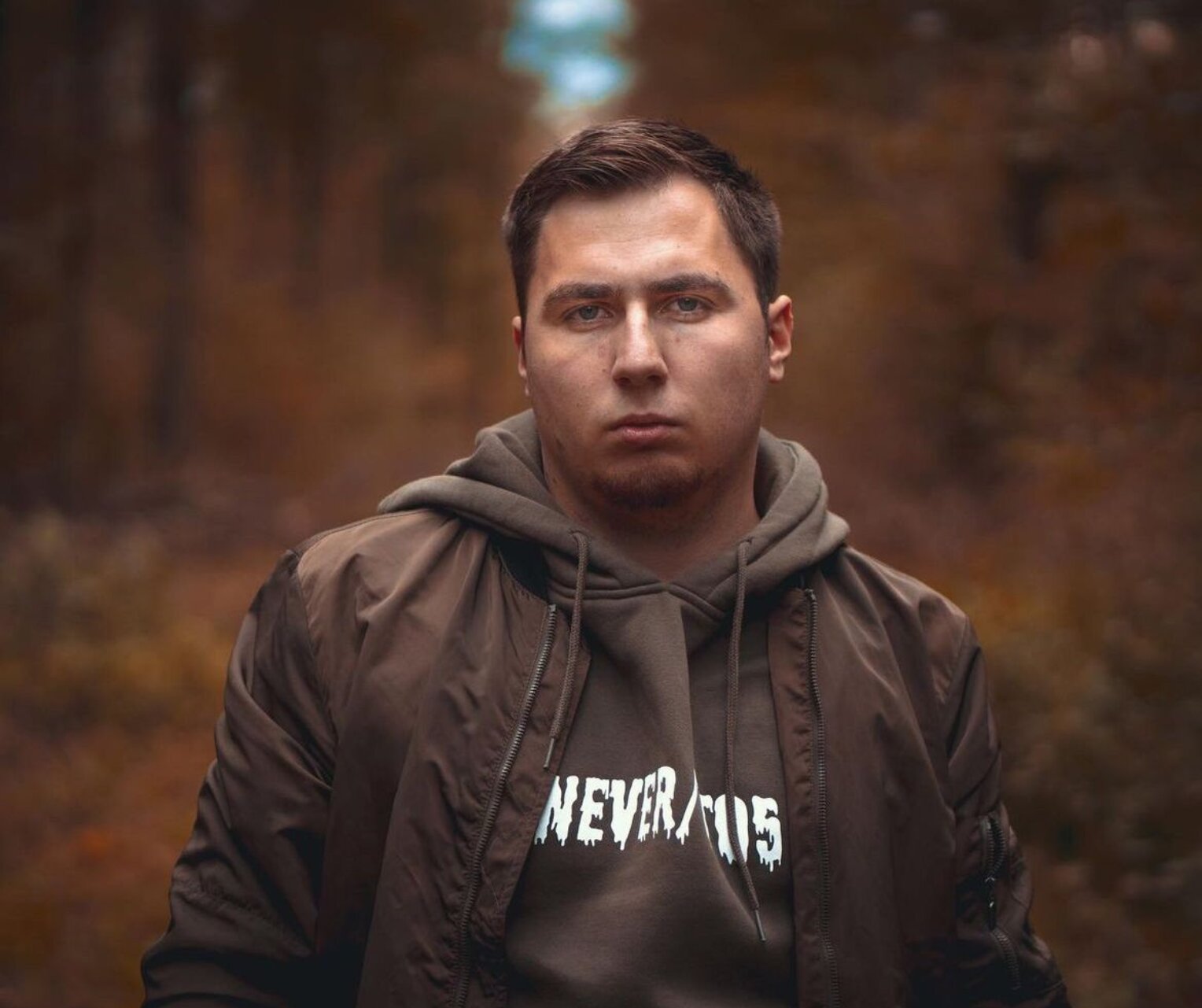Porträt eines jungen Mannes mit ernstem Blick, der in einem herbstlichen Wald steht. Er trägt eine braune Bomberjacke und einen Hoodie mit dem Aufdruck „NEVER TOO“.