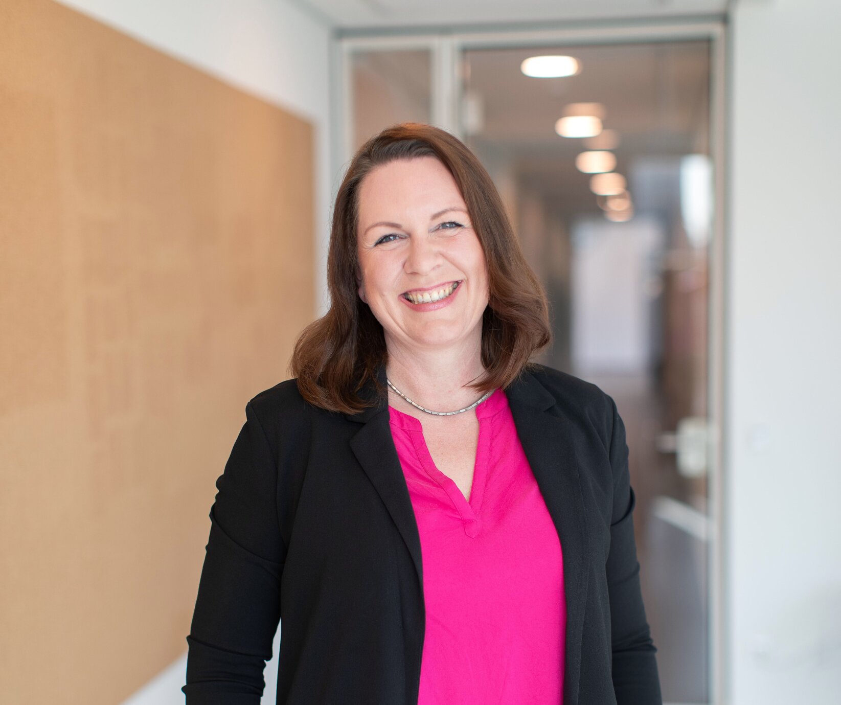Porträt einer lächelnden Frau in einem modernen Büroflur. Sie trägt eine pinke Bluse und einen schwarzen Blazer.