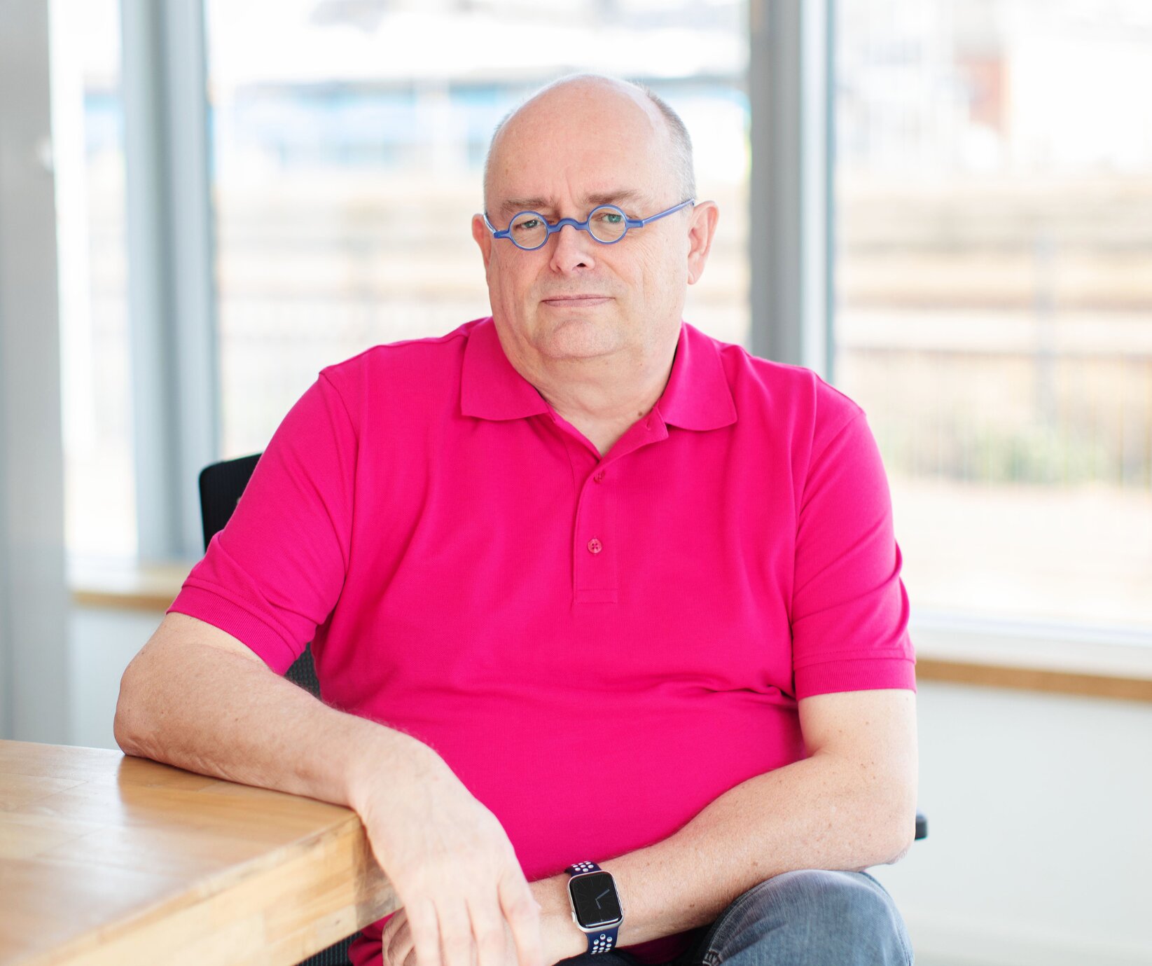 Porträt eines Mannes mit pinkem Poloshirt und blauer Brille, sitzend an einem Holztisch in einem lichtdurchfluteten Büro.